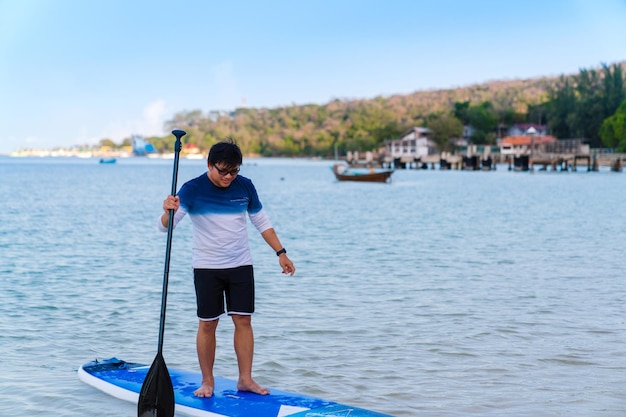 Homem asiático com óculos em roupas casuais, em pé, stand up paddleboard, homem feliz paddle boarding na água do mar azul nas férias de verão na praia da ilha da tailândia