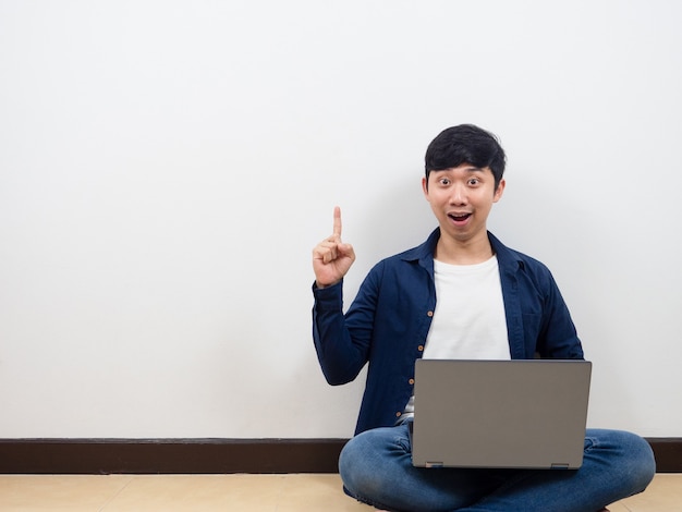 Homem asiático com seu laptop sentado no chão levanta o dedo indicador sobre o fundo da parede branca