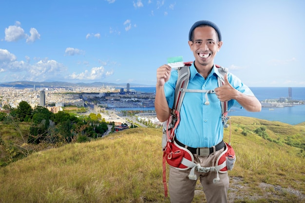 Homem asiático com um gorro e uma mochila segurando cartão com um polegar para cima gesto em pé na colina