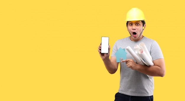 Homem asiático de arquiteto Choque e surpresa de segurar a forma de papel em casa, apresentando o telefone inteligente isolado na parede amarela