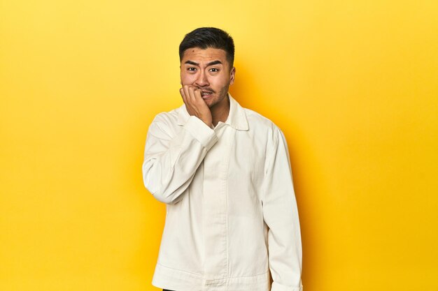 Foto homem asiático de camisa branca no estúdio amarelo a morder as unhas nervoso e muito ansioso