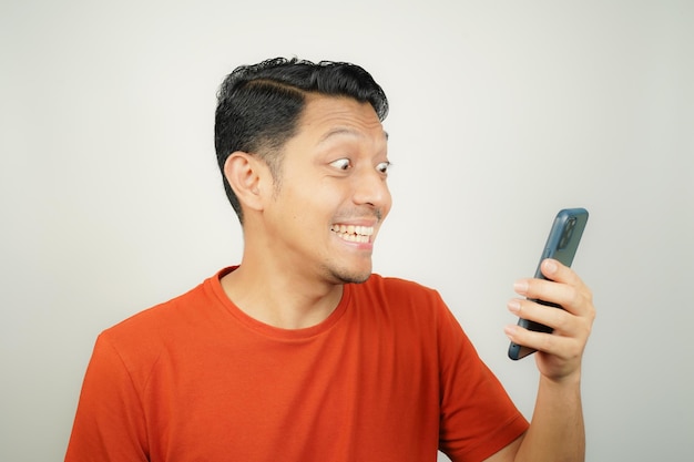 Homem asiático de camiseta laranja feliz olhando para o que no smartphone em fundo isolado