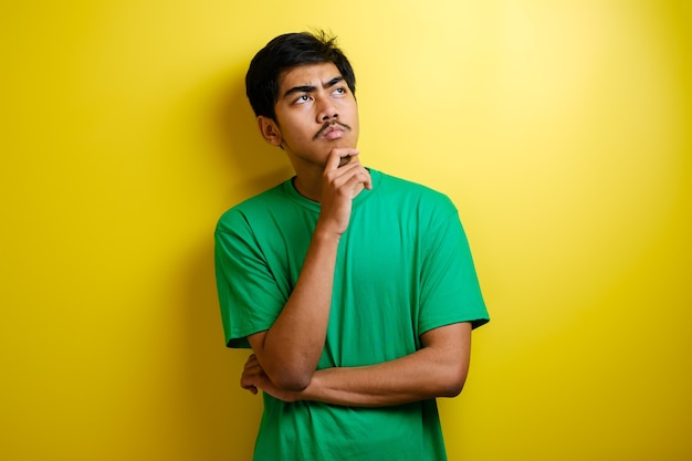 Homem asiático de camiseta verde pensando em alguma coisa, procurando uma boa ideia contra um fundo amarelo