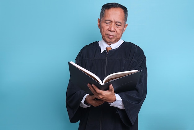 Homem asiático de juiz sênior em um vestido carregando um livro de direito isolado no fundo azul