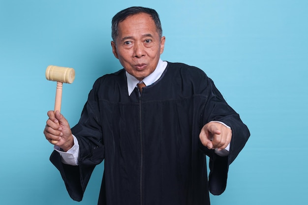 Homem asiático de juiz sênior surpreso e apontando para frente isolado em fundo azul