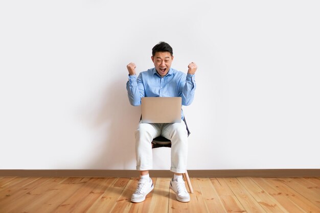 Homem asiático de meia-idade muito feliz com computador portátil sentado na cadeira comemorando muito fazendo gesto SIM