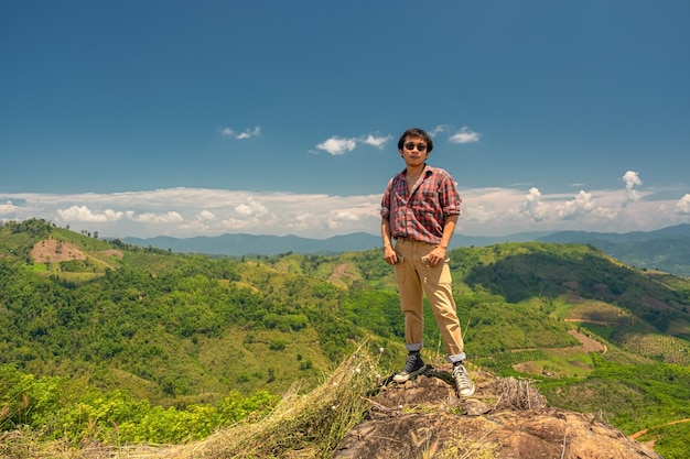 Homem asiático de pé em uma rocha com o céu e as montanhas ao fundo