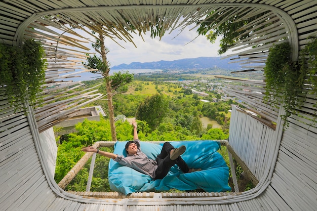 Homem asiático deitado no sofá-cama homem turista asiático está deitado no sofá-cama perto do terraço no café ao ar livre com vista panorâmica sobre as montanhas. hora de relaxar