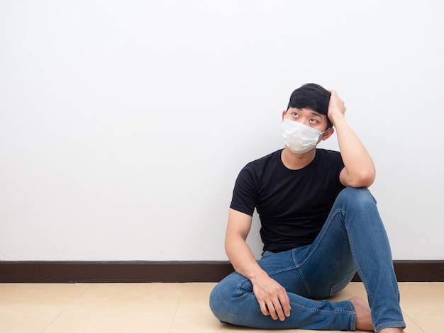 Homem asiático deprimido usando máscara sentado no chão tocando sua cabeça e olhando para um fundo branco