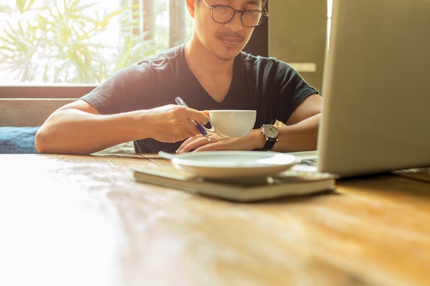 Homem asiático do retrato com os vidros que têm a ruptura de café que trabalha com portátil.