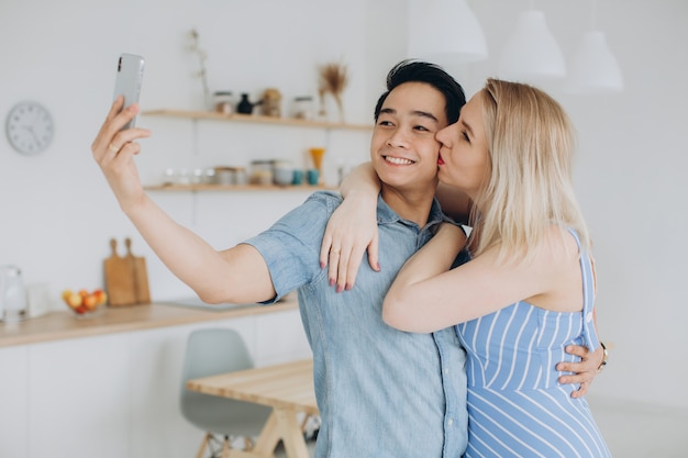 Homem asiático e sua mulher loira caucasiana passam tempo juntos e fazendo selfie um para o outro na cozinha