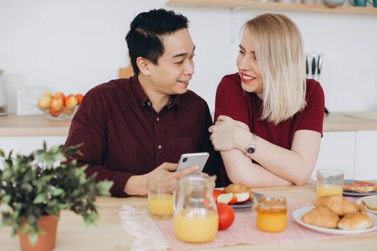 Foto homem asiático e sua mulher loira caucasiana tomar café da manhã. eles passam um tempo juntos na cozinha e olham o vídeo no telefone.