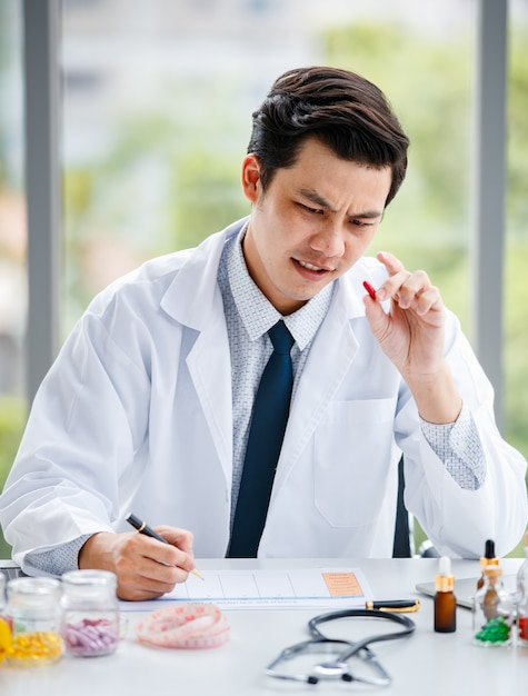 Homem asiático em uniforme médico mostrando a pílula e fazendo anotações enquanto trabalhava em uma clínica moderna