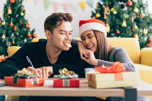 Homem asiático escrevendo em um cartão postal de Natal em branco com uma caneta enquanto sua namorada lhe dá uma batata frita Casal sentado e escrevendo cartão de Natal juntos em casa durante o feriado de Natal