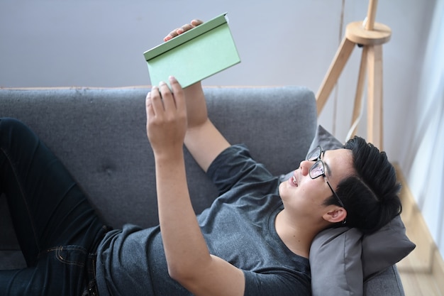 Homem asiático feliz deitado no sofá confortável e lendo o livro.