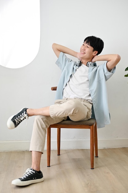 Homem asiático feliz em roupas casuais senta-se em uma cadeira relaxando em sua sala de estar brilhante mínima