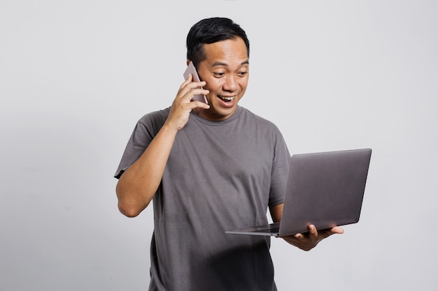 Homem asiático feliz fazendo uma ligação usando um telefone inteligente, segurando e olhando para um laptop