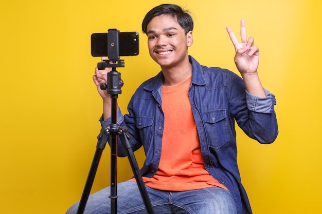 Homem asiático feliz gravando vídeo mostrando vsign isolado sobre fundo amarelo brilhante