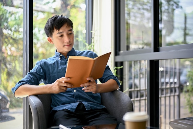 Homem asiático feliz lendo um livro enquanto relaxa em uma cafeteria