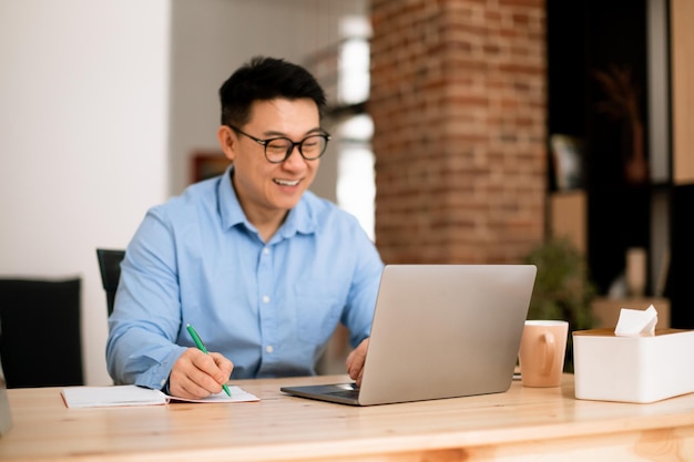 Homem asiático feliz olhando para a tela do laptop e escrevendo no notebook sentado no local de trabalho no foco seletivo do home office