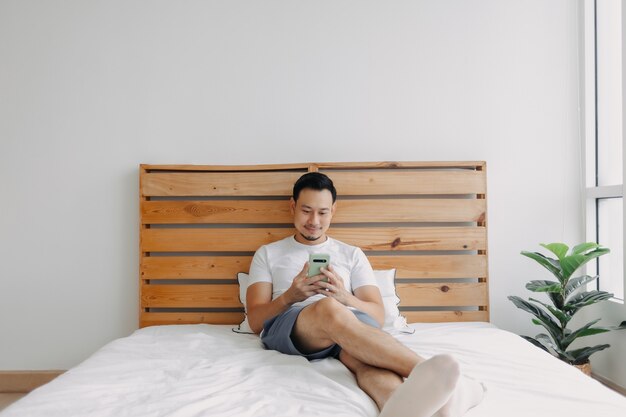 Homem asiático feliz relaxando com o smartphone na cama