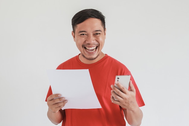 Homem asiático feliz vestindo camiseta vermelha, olhando a câmera enquanto segura um papel branco em branco e um smartphone