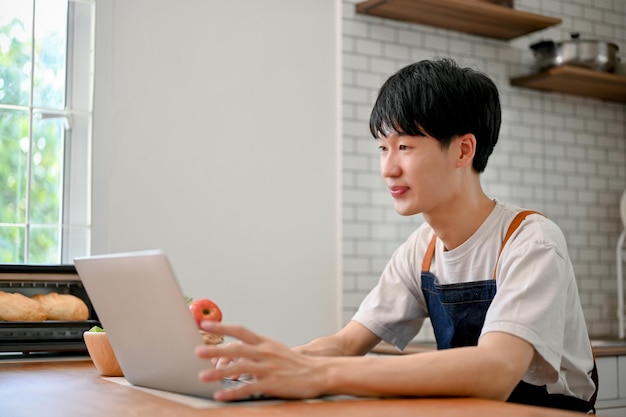 Homem asiático focado em avental usando laptop em sua cozinha participando de aula de culinária online