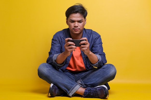 Homem asiático focado em roupas casuais jogando jogos no smartphone sentado de pernas cruzadas isoladas sobre