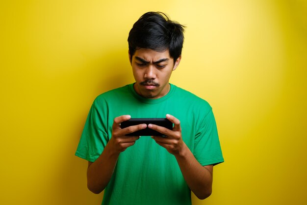 Homem asiático joga jogo para celular em seu smartphone com uma expressão séria ou zangada contra um fundo amarelo