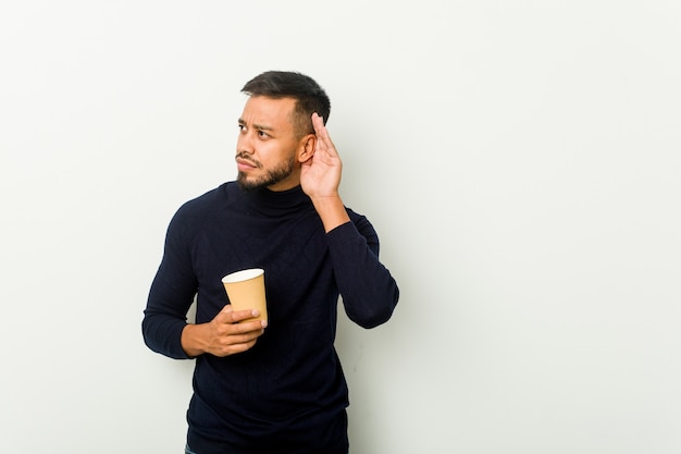 Homem asiático jovem de raça mista segurando um café para viagem, tentando ouvir uma fofoca.