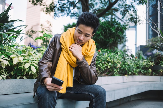 Homem asiático mensagens no smartphone enquanto está sentado no banco de pedra