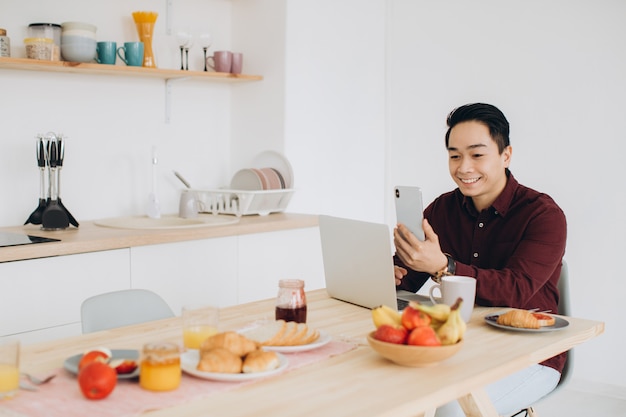 Homem asiático moderno trabalhando no laptop na cozinha no café da manhã.