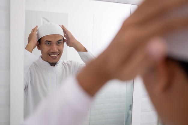 Homem asiático muçulmano olhando para o espelho e se vestindo antes de ir para a mesquita usando um boné ou chapéu islâmico