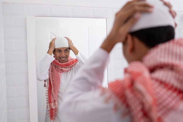 Homem asiático muçulmano olhando para o espelho e se vestindo antes de ir para a mesquita usando um boné ou chapéu islâmico