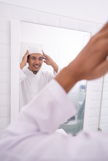 Homem asiático muçulmano olhando para o espelho e se vestindo antes de ir para a mesquita usando um boné ou chapéu islâmico