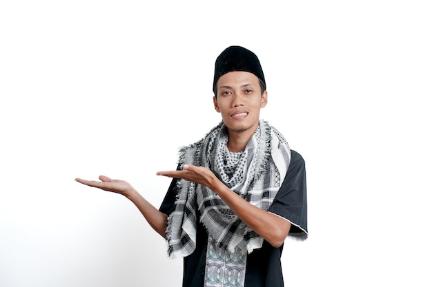 Homem asiático muçulmano religioso usando vestido muçulmano turbante e boné apontando para o lado do espaço vazio isolado no fundo branco
