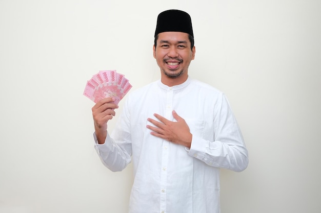 Homem asiático muçulmano sorrindo e mostrando gesto grato enquanto segura dinheiro