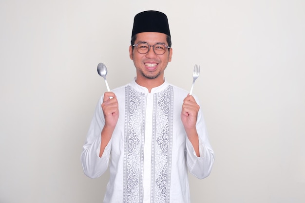 Homem asiático muçulmano sorrindo feliz com a mão segurando colher e garfo