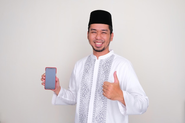 Homem asiático muçulmano sorrindo feliz e desistindo do polegar enquanto segura a tela do telefone em branco