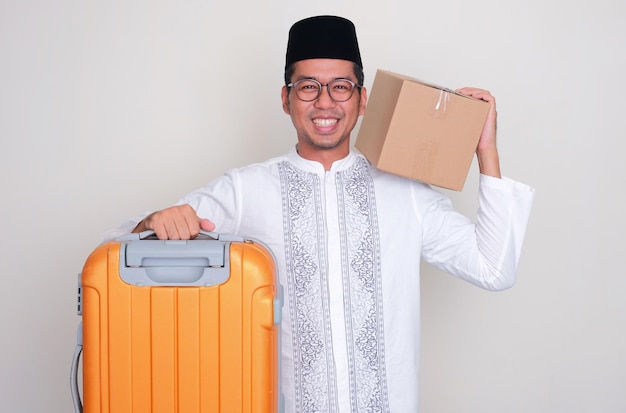 Homem asiático muçulmano sorrindo feliz enquanto carrega suticase e caixa de pacote