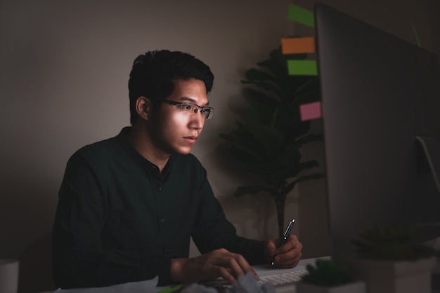 Homem asiático novo atrativo que senta-se na tabela da mesa que olha o laptop no trabalho tarde da noite escuro que sente o pensamento sério e o escritório determinado em casa no trabalho duro ou no conceito de trabalho da carga de trabalho.