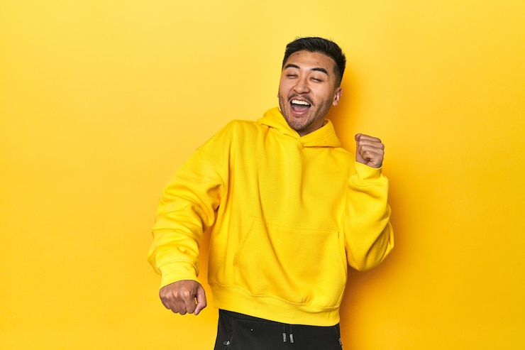 Foto homem asiático ousado com capuz amarelo em um estúdio amarelo dançando e se divertindo