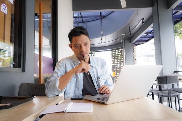 Homem asiático pensa e procura ideias com camisa azul usando laptop e telefone celular em uma cafeteria