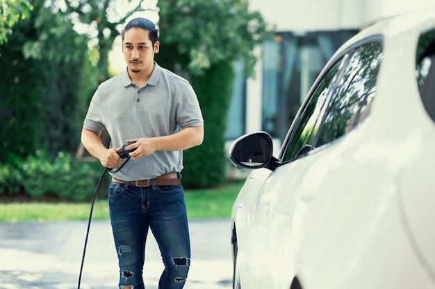 Homem asiático progressivo recarrega seu carro EV na estação de carregamento em casa