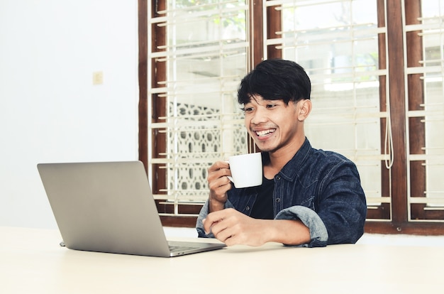 Homem asiático sentado em frente a um laptop tomando café