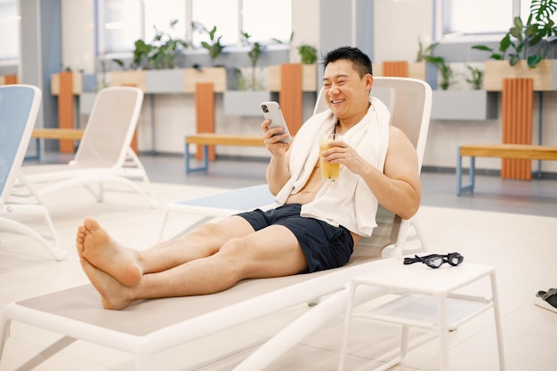 Homem asiático sentado em uma espreguiçadeira na piscina interna e usando um telefone