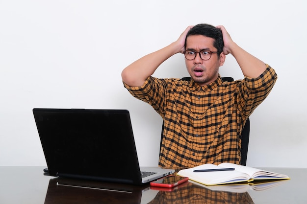 Homem asiático sentado na mesa de trabalho mostrando expressão chocada ao olhar para seu laptop