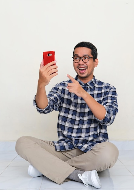 Homem asiático sentado no chão olhando e apontando para seu handphone