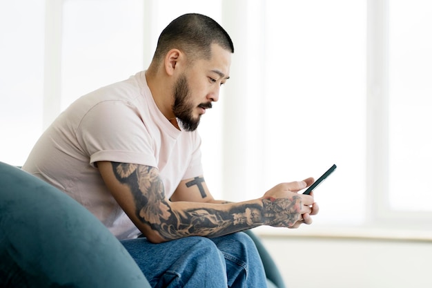 Homem asiático sério e bonito usando telefone celular, fazendo compras on-line sentado em casa. Tecnologia