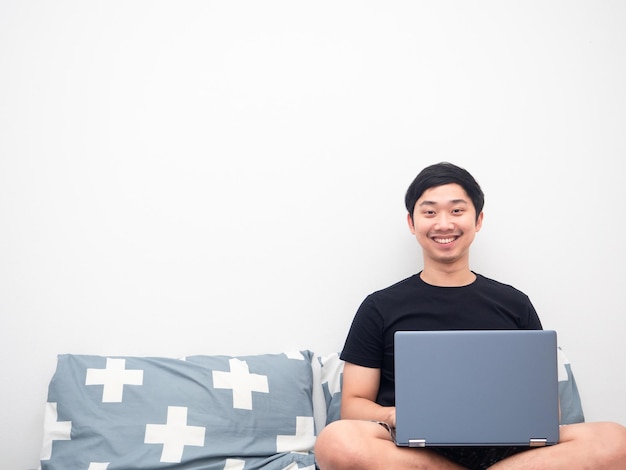 Homem asiático sorri sentado na cama com espaço de cópia de fundo branco do laptop